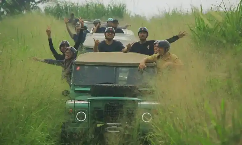 Peserta wisata petualangan sedang menikmati perjalanan offroad menggunakan mobil Jeep Land Rover terbuka di jalur berbatu dan berumput Bogor bersama provider Paintball Bogor.