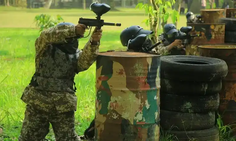 Pemain Paintball Bogor dalam posisi taktis membidik dari balik barikade tong dan ban bekas, mengenakan seragam camouflage digital lengkap dan masker keselamatan di medan outbound.
